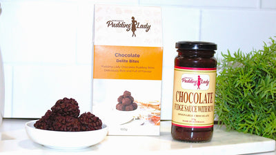 Jar of chocolate fudge sauce with rum, a box of chocolate delite bites, and a bowl of chocolate delite bites on a white surface with a plant in the background.