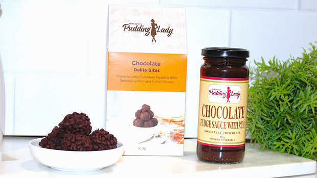Jar of chocolate fudge sauce with rum, a box of chocolate delite bites, and a bowl of chocolate delite bites on a white surface with a plant in the background.