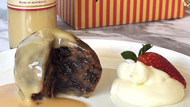 Dessert with a slice of pudding, cream, and strawberry on a plate, with brandy custard and gift box in background