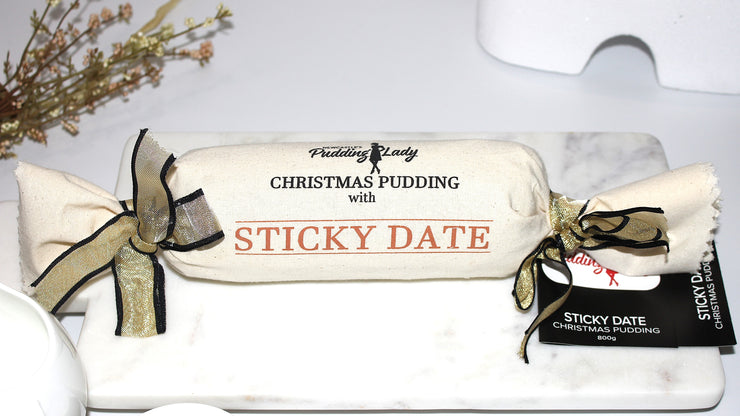 Christmas pudding with sticky date, wrapped in white calico with gold ribbon, on a white plate.