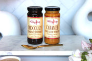 Two jars of Pudding Lady fudge sauce, one chocolate and one caramel, on a white surface with a spoon and flowers.