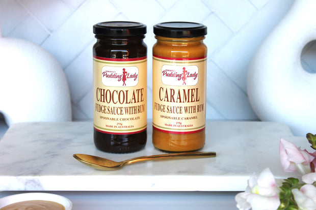 Two jars of Pudding Lady fudge sauce, one chocolate and one caramel, on a white surface with a spoon and flowers.