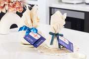 Two wrapped gluten free puddings with ribbons and tags on a table with flowers in the background