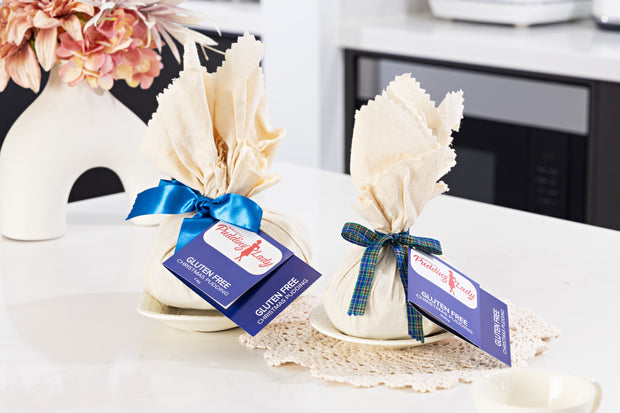 Two wrapped gluten free puddings with ribbons and tags on a table with flowers in the background