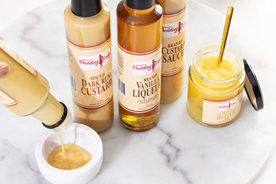 Pudding Lady sweet sauces including bottle of Spiced Dark Rum Custard on a marble surface.