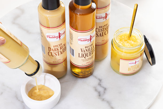 Pudding Lady sweet sauces including bottle of Spiced Dark Rum Custard on a marble surface.