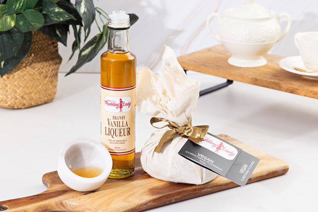 Bottle of vanilla liqueur on a wooden board with a wrapped Pudding Lady vegan pudding. 