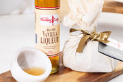 Bottle of vanilla liqueur on a wooden board with a wrapped Pudding Lady vegan pudding. 