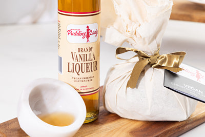 Bottle of vanilla liqueur on a wooden board with a wrapped Pudding Lady vegan pudding. 